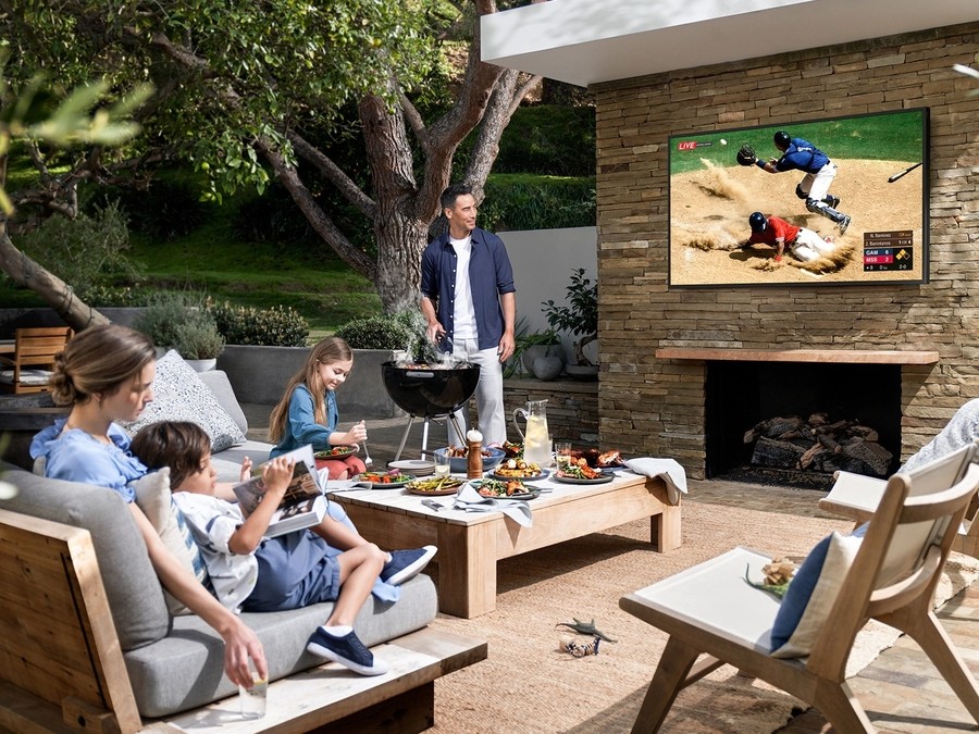 A man grills while the whole family enjoys a ballgame on an outdoor TV mounted over a brick patio fireplace.